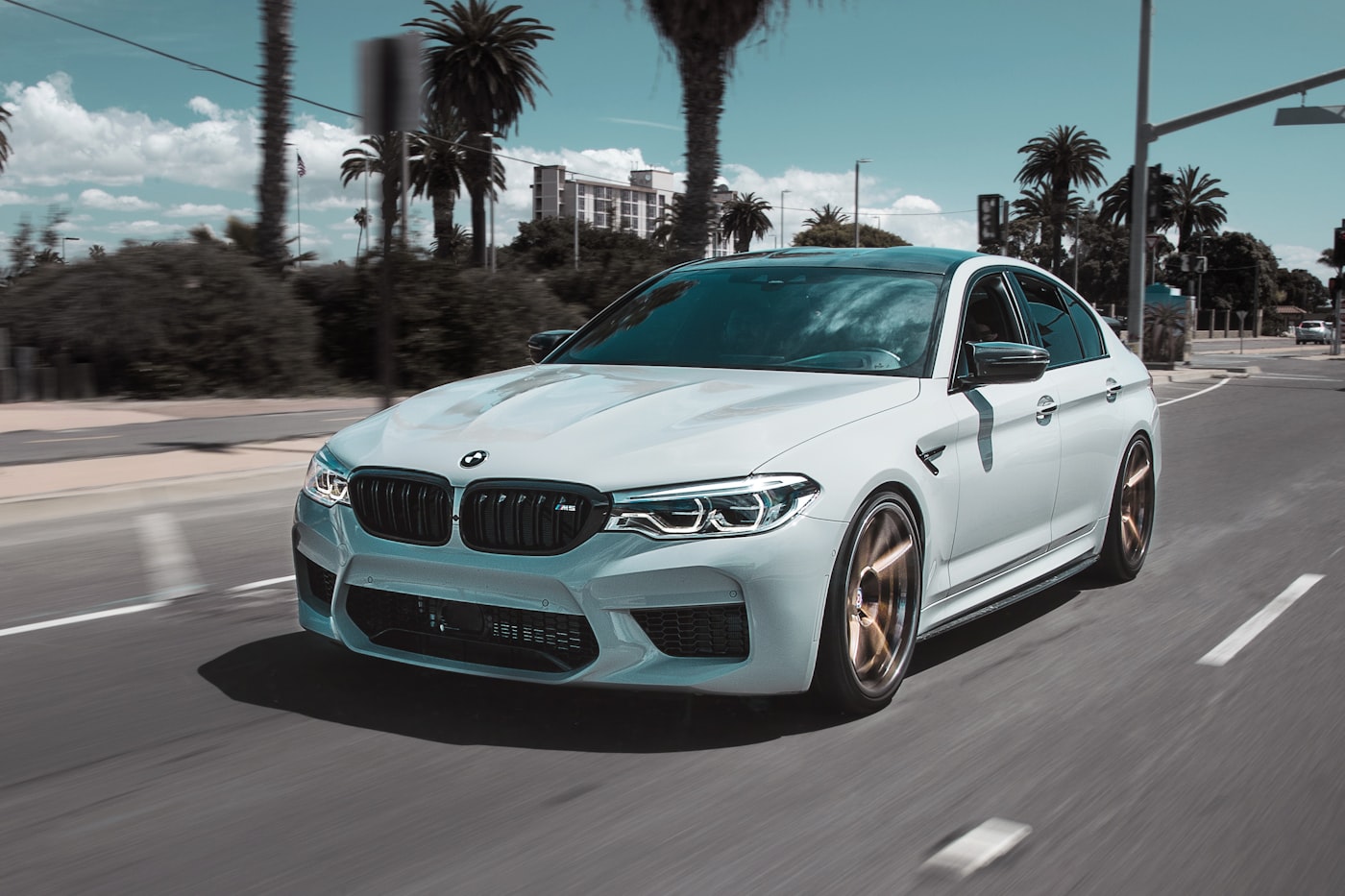 BMW M5 blanco en movimiento por una avenida con palmeras.